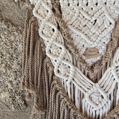Tapeçaria de macramé bege e branca com padrão geométrico pendurada em madeira