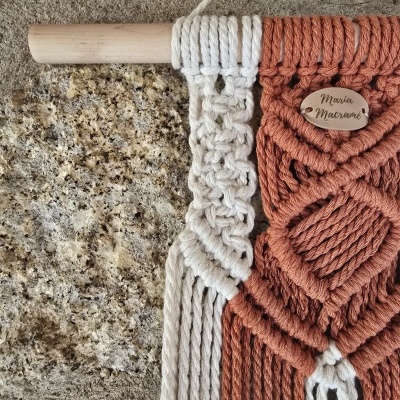 Tapeçaria de macramé branca e terracota com etiqueta de madeira pendurada num suporte de madeira num fundo de pedra