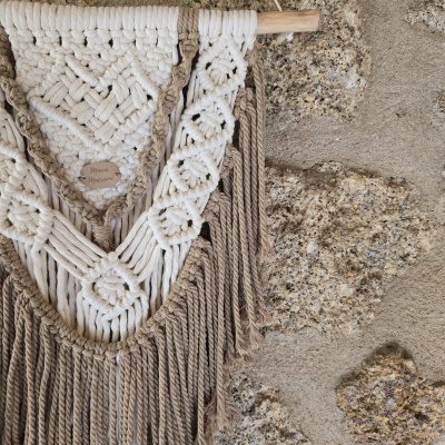 Tapete de macramé bege e castanho pendurado em parede de pedra com etiqueta Boho Macramé