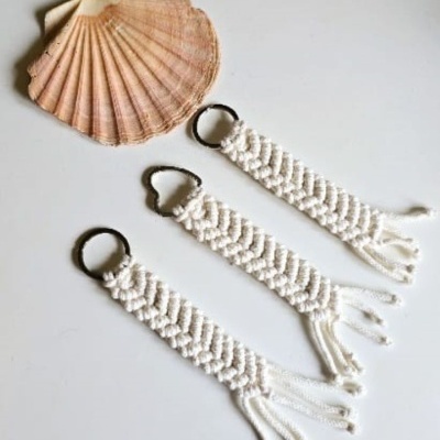 Três porta-chaves de macramé bege com anéis metálicos escuros ao lado de conchas sobre superfície branca