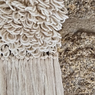 Pano decorativo em macramé creme com nós e franjas pendurado numa parede de pedra rústica.
