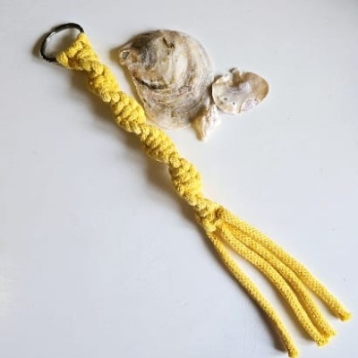 Chaveiro amarelo em macramé com argola preta e franjas, ao lado de conchas sobre fundo branco.