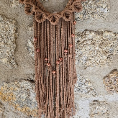 Painel decorativo de macramé castanho com franjas e contas de madeira pendurado numa vara de madeira contra parede de pedra