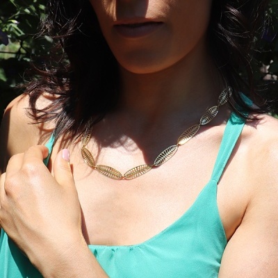 Colar dourado com elementos ovais usados em pescoço de mulher com vestido verde-água ao ar livre
