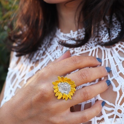 Anel de girassol amarelo e prateado numa mão feminina com unhas azuis sobre blusa branca de renda.