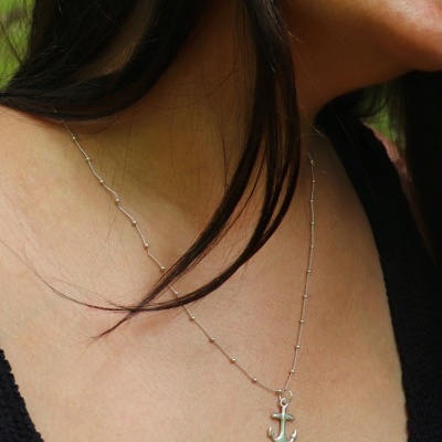 Colar prateado com pendente âncora no pescoço de mulher com cabelo castanho e roupa preta