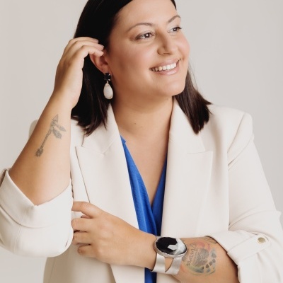 Mulher com blazer bege e acessórios vistosos em fundo neutro