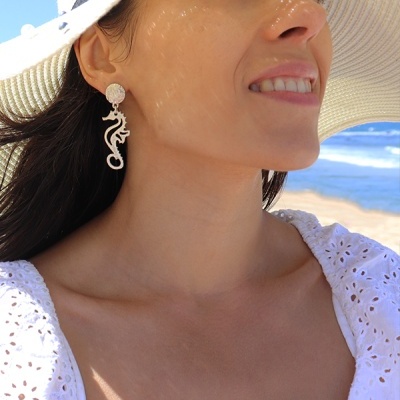 Brincos cavalo-marinho prateados usados por mulher com chapéu e vestido branco na praia.