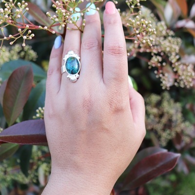 Mão com anel de prata com pedra azul esverdeada entre folhas e botões florais
