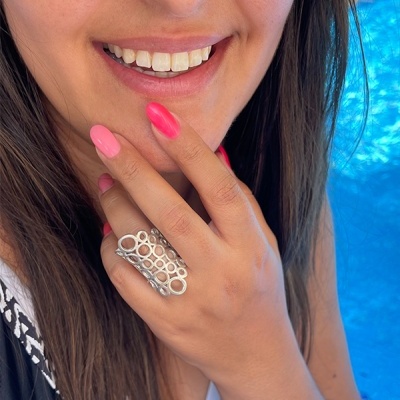 Mão feminina com anel prateado e unhas rosas junto ao rosto sorridente