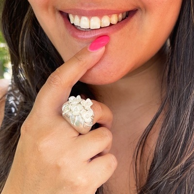 Anel prateado de flores num dedo indicador com unhas rosas perto da boca sorridente