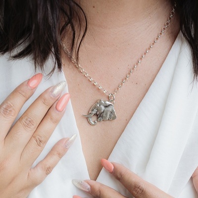 Colar prateado com pendente de elefante visto no pescoço de uma mulher com blusa branca e unhas pintadas