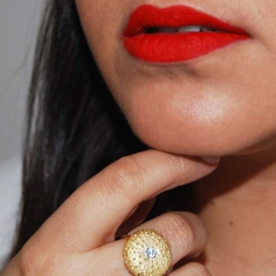 Mão com anel dourado texturizado e pedra brilhante, rosto com batom vermelho.