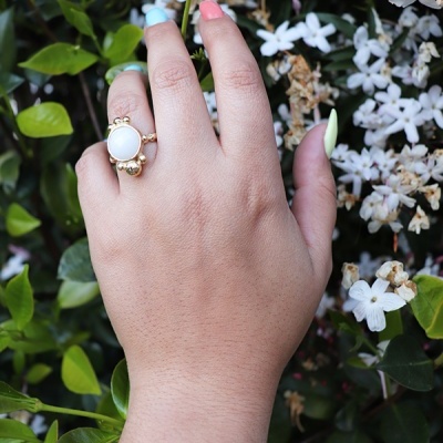 Mão com anel dourado e pedra branca entre flores brancas e folhas verdes