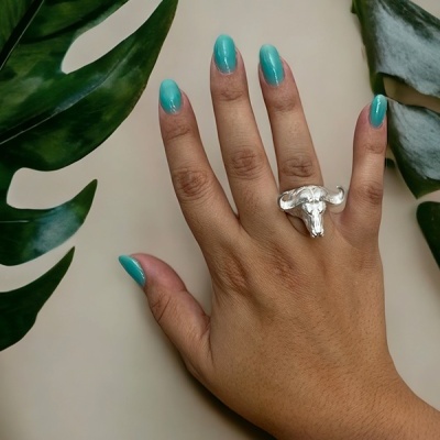 Mão feminina com anel prateado em forma de cabeça de búfalo e unhas pintadas de azul turquesa, com folhas verdes ao redor.