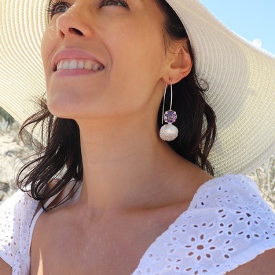 Brincos pendentes com pedra roxa e pendente branco texturizado em mulher com chapéu branco e vestido branco na praia
