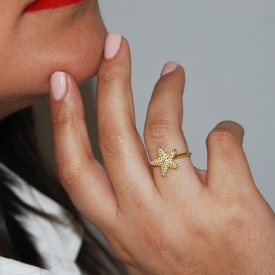 Anel dourado de estrela do mar numa mão feminina com unhas rosas e batom vermelho