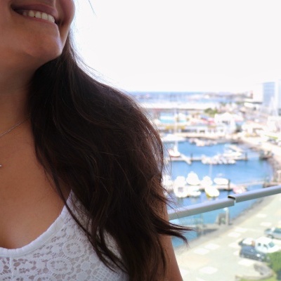 Colar prateado com estrela-do-mar e pedra redonda branca num pescoço feminino com blusa branca de renda, com marina ao fundo.