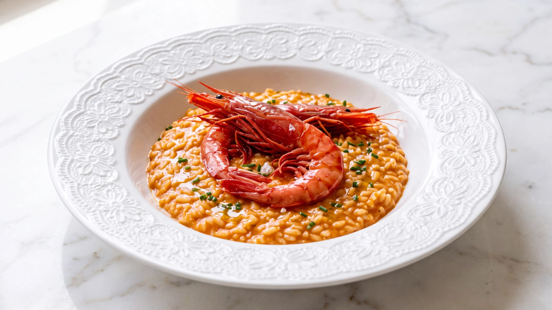 ARROZ MELOSO DE CARABINEIRO Prato com risoto cremoso alaranjado e dois camarões vermelhos no centro num prato branco com padrão floral.
