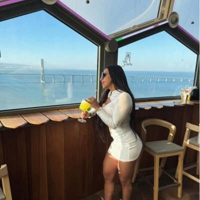 Mulher com vestido branco e óculos escuros segurando copo com bebida amarela junto a janela com vista para ponte