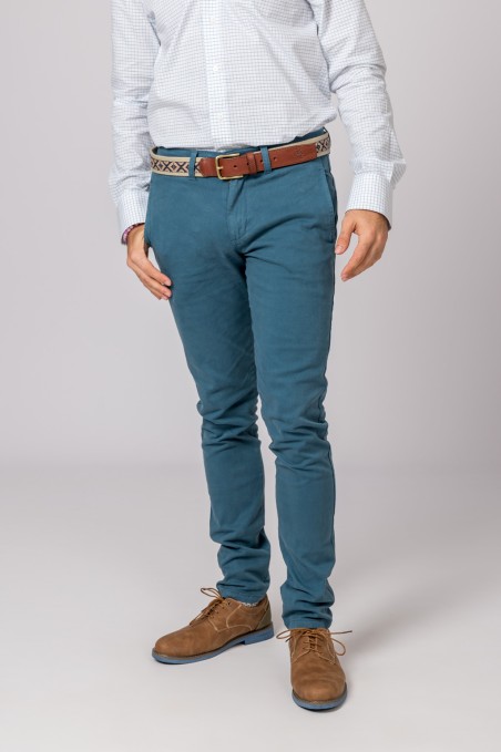 Homem com camisa branca, calças azuis, cinto castanho e sapatos castanhos em fundo neutro