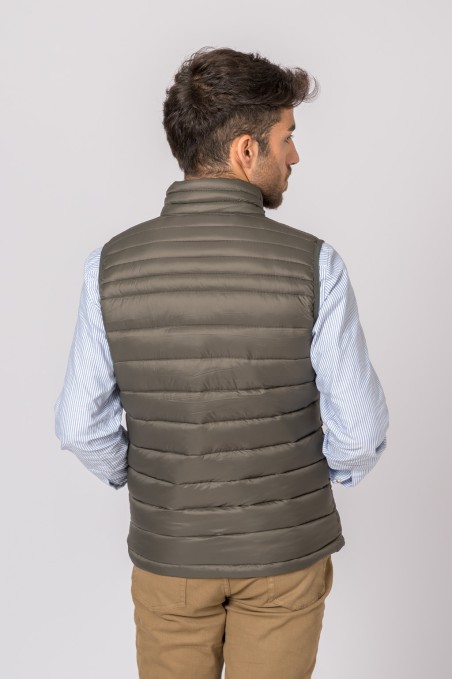 Homem de costas com colete verde-oliva acolchoado sobre camisa azul clara e calças bege