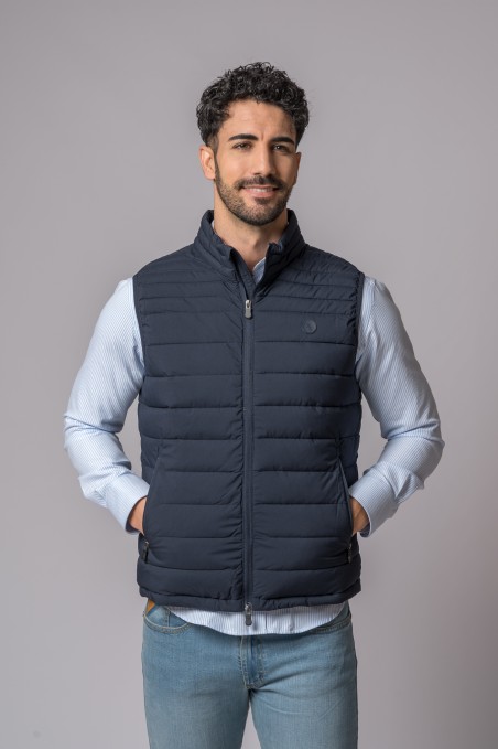 Homem a usar colete acolchoado azul escuro com camisa azul clara e jeans em fundo cinzento