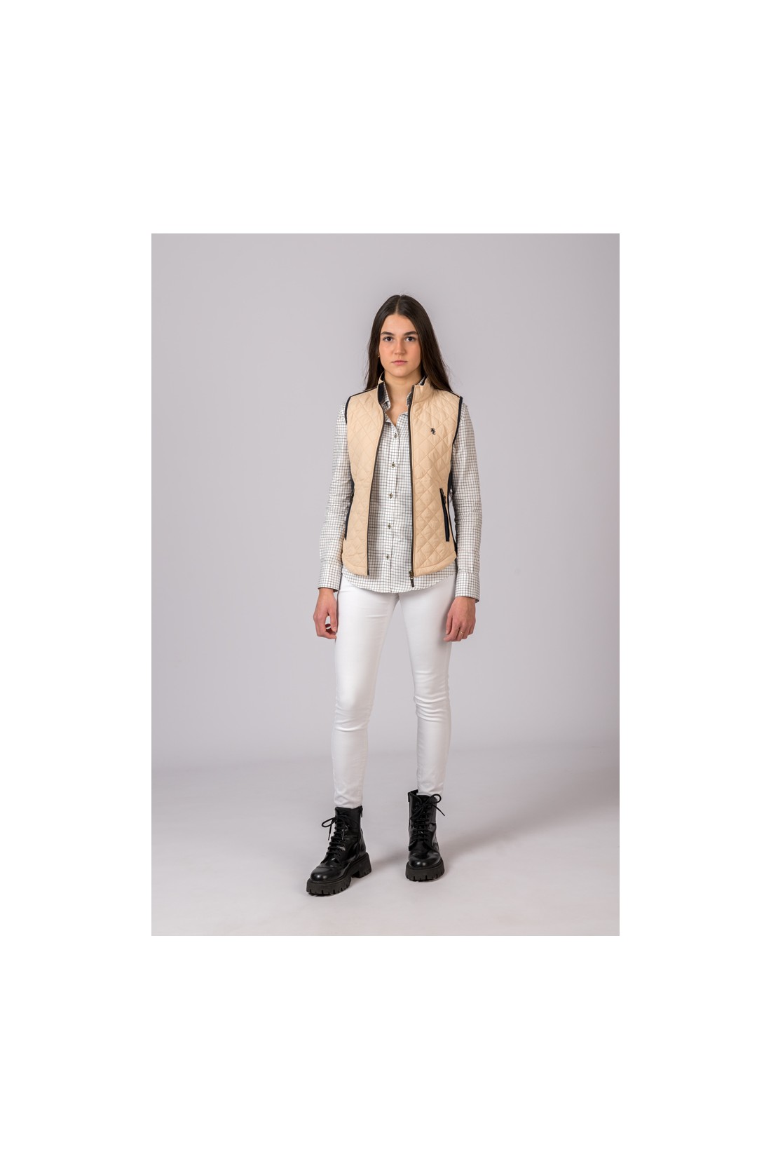 Mulher com colete bege, camisa cinza às riscas, calças brancas e botas pretas