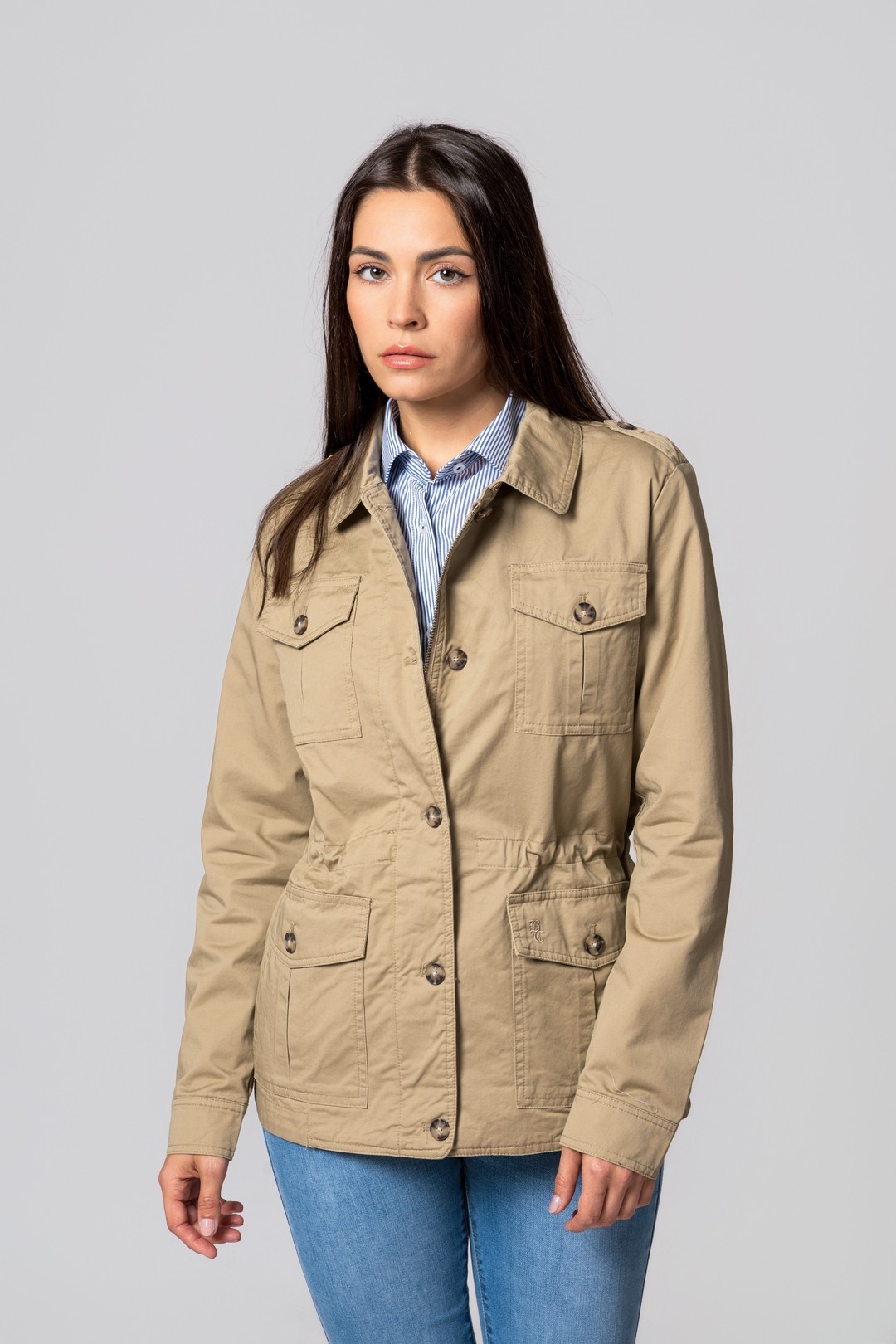 Casaco bege claro com botões e bolsos, mulher com camisa riscada e jeans, fundo cinza claro