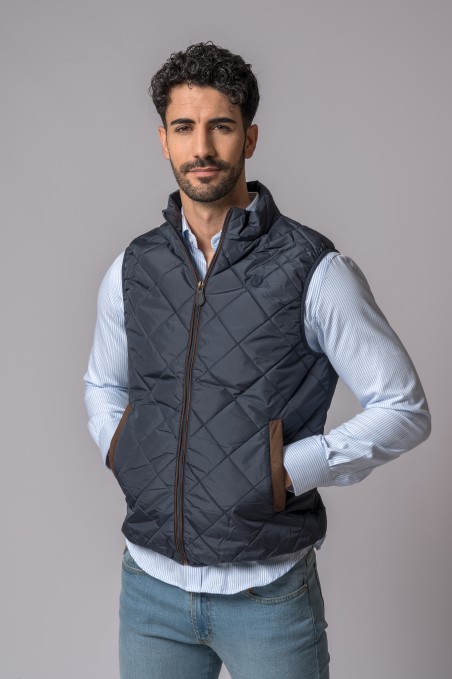 Colete acolchoado preto com bolsos castanhos usado sobre camisa azul com riscas finas e jeans azuis