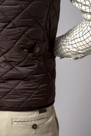 Colete acolchoado castanho com padrão em losangos, usado com camisa xadrez clara e calças bege