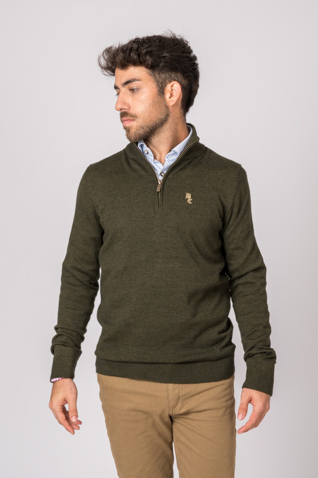 Camisola verde escura de malha com gola subida e fecho, camisa azul clara e calças bege, homem em fundo cinzento claro.