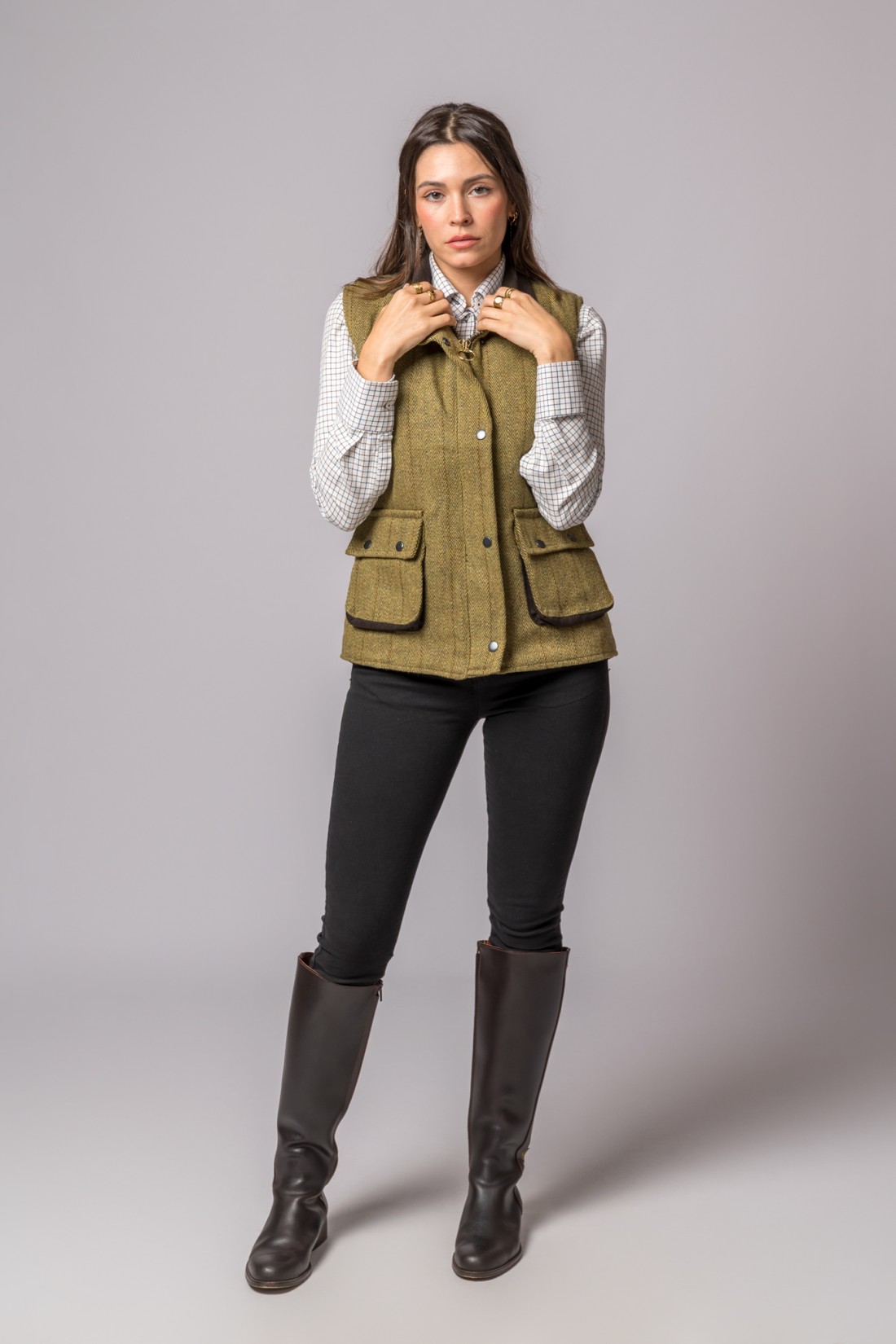 Mulher vestindo colete verde oliva com botões, camisa xadrez branca e calças pretas com botas castanhas.