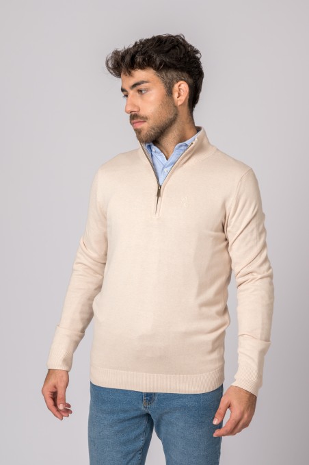 Homem com camisola bege de gola alta e fecho, camisa azul clara e calças de ganga azuis