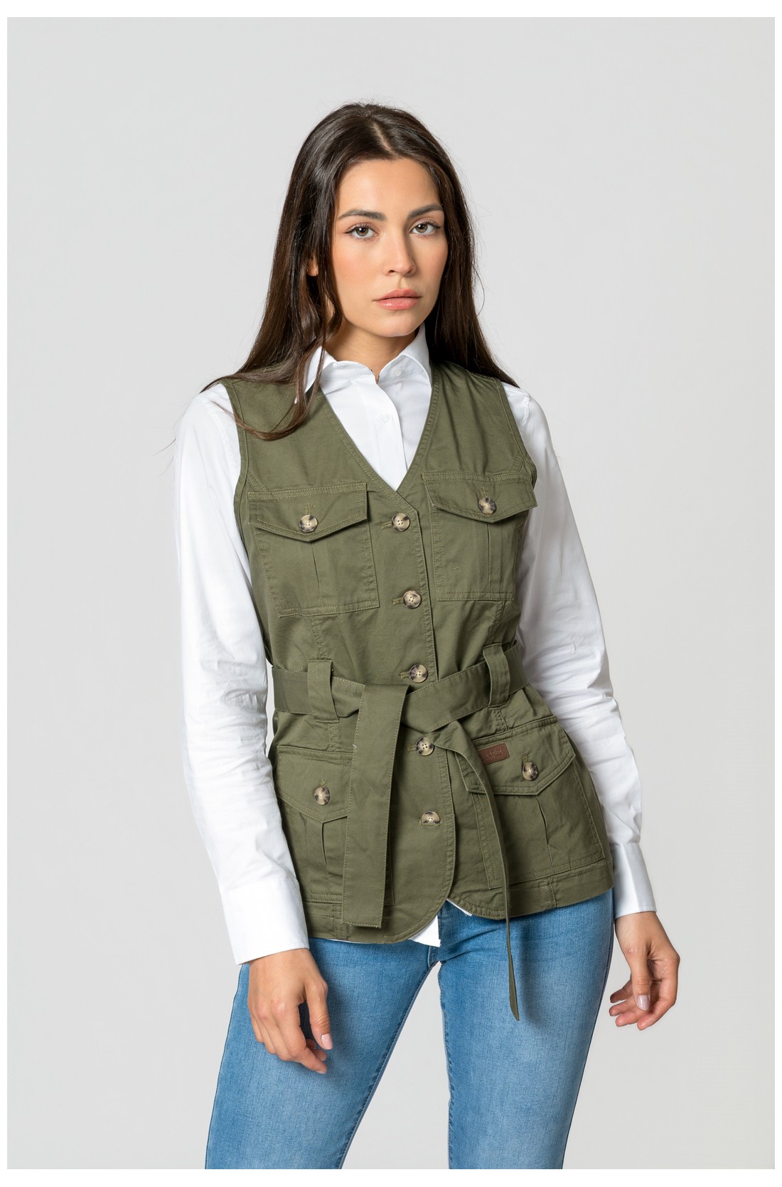 Colete verde caqui com cinto e botões usado sobre camisa branca e jeans azul claro