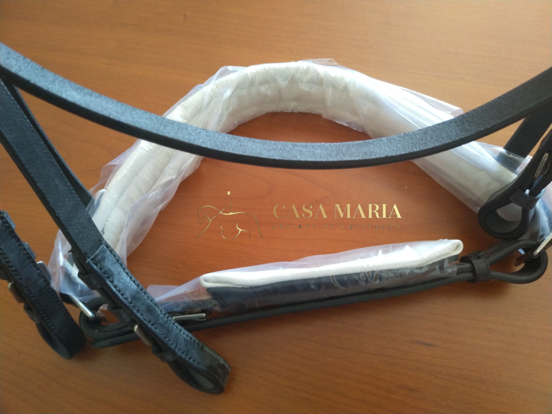 Arreios para cavalo em couro preto protegidos parcialmente por plástico sobre mesa de madeira com texto CASA MARIA