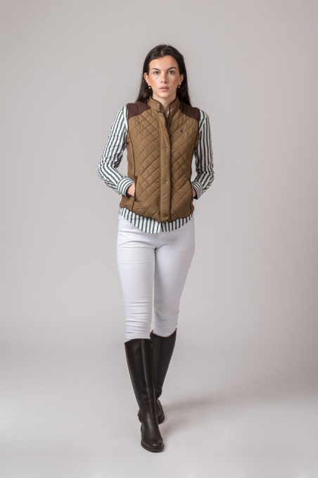 mulher de pé com colete acolchoado castanho, camisa de riscas, calças brancas e botas pretas
