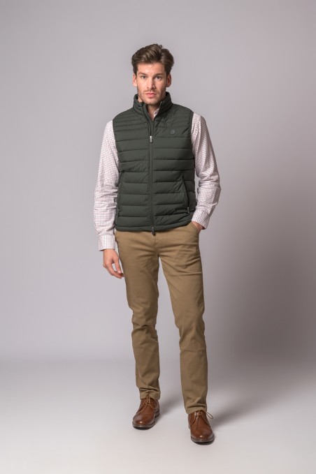 Homem com colete acolchoado verde, camisa xadrez, calças bege e sapatos castanhos em fundo cinzento