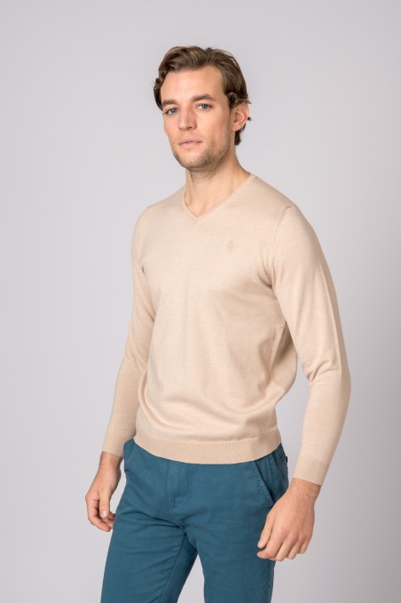 Homem com camisola bege de manga comprida e calças azuis sobre fundo cinzento