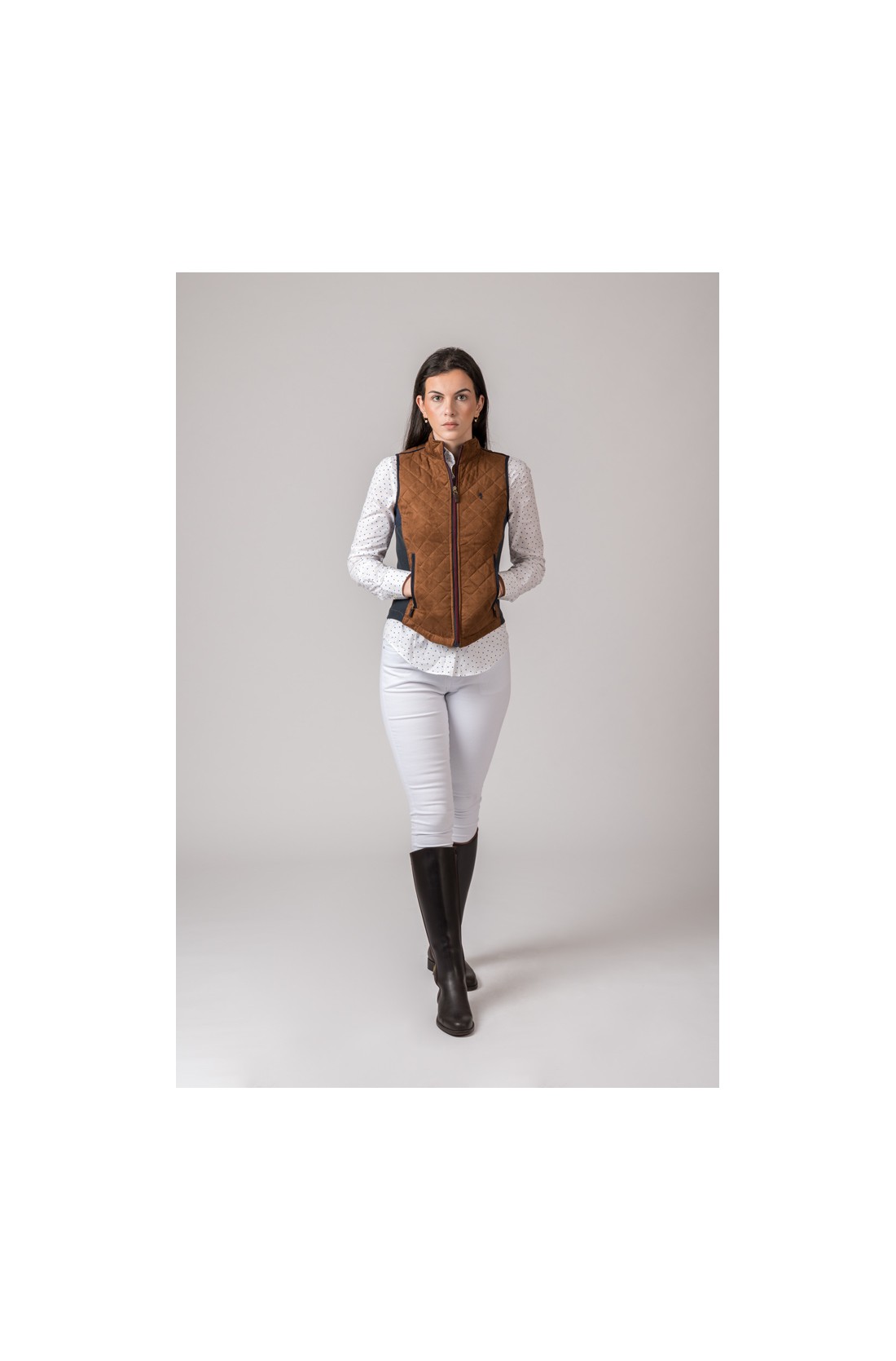 Mulher com colete castanho acolchoado, camisa branca de pontos, calças brancas e botas pretas
