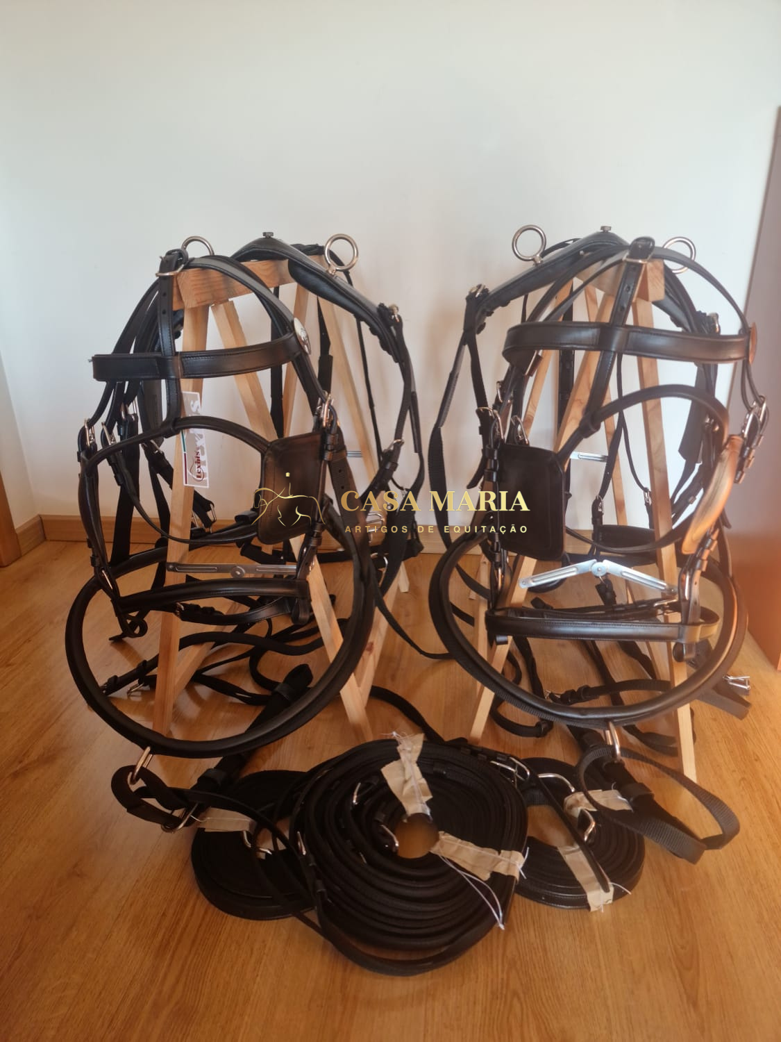 Dois arreios de cavalo em couro preto com rédeas, em cavaletes de madeira, sobre piso de madeira, fundo branco