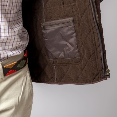 Interior de casaco castanho com bolso interno com fecho, usado por pessoa com camisa xadrez branca e calças claras.