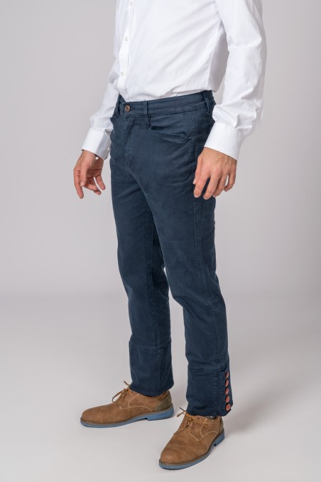 Calças azuis escuras de homem com detalhes vermelhos na bainha, camisa branca e sapatos castanhos em fundo cinzento