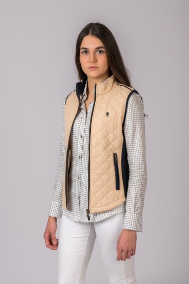 Mulher com colete bege acolchoado com detalhes pretos, camisa xadrez branca e preta e calças brancas