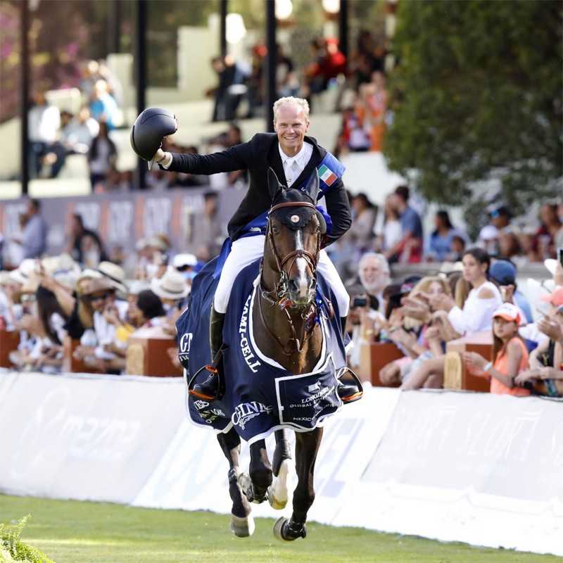 Cavaleiro vestido formal em cavalo com manta azul e logos brancos, em evento ao ar livre com público