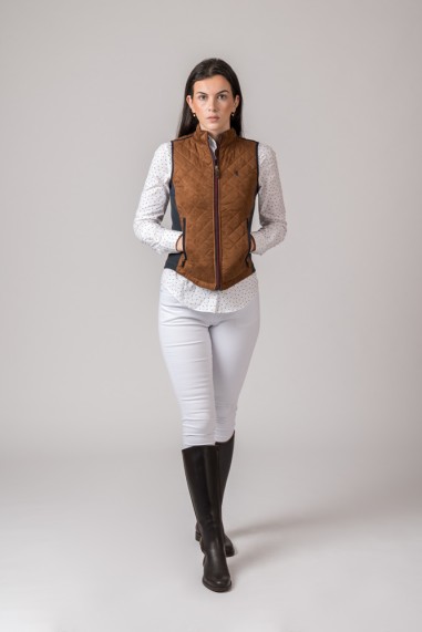 Mulher com colete castanho acolchoado e botas pretas em fundo cinzento claro