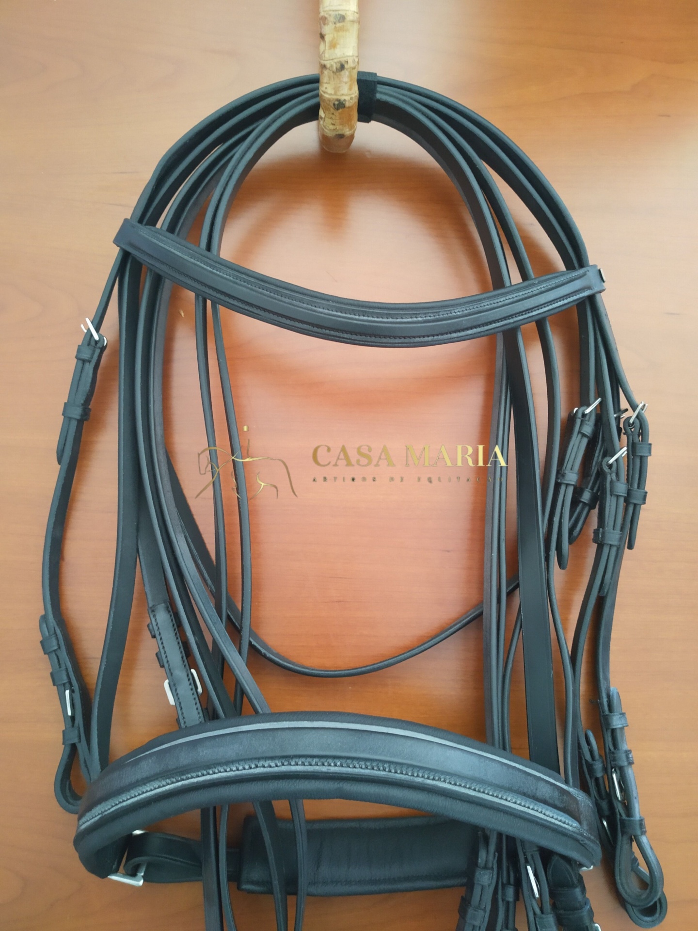 Bridão preto de couro com fivelas metálicas sobre madeira clara com texto CASA MARIA