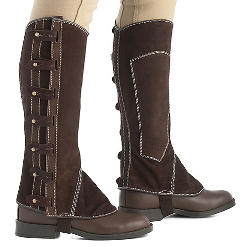 Perneiras de equitação castanhas com correias laterais usadas com calças claras.