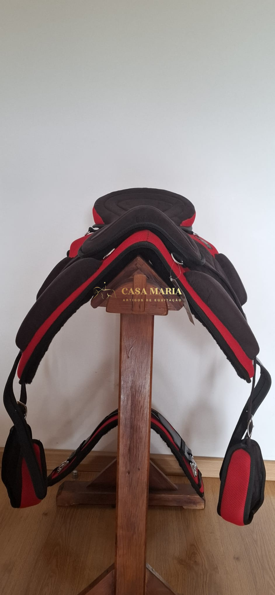 Selim de equitação preto e vermelho pendurado em suporte de madeira com texto CASA MARIA