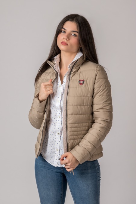 Mulher a usar casaco bege acolchoado com emblema no peito, camisa branca com padrões e jeans azuis